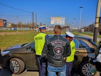 Na zdjęciu widoczny jest motocyklista ubrany w skórzaną kurtkę. Stoi tyłem do obiektywu kamery. Przed nim znajdują się dwaj policjanci Wydziału Ruchu Drogowego ubrani w odblaskowe kamizelki stojący przy swoim radiowozie. Policjanci odwróceni są tyłem do obiektywu aparatu.