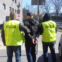 MĘŻCZYZNA W CZARNEJ CZAPCE Z DASZKIEM W KAJDANKACH PROWADZONY PRZEZ POLICJANTÓW W KAMIZELKACH ODBLASKOWYCH Z NAPISEM POLICJA. W TLE WIDOCZNY PARKING SAMOCHODOWY ORAZ BUDYNEK I KOMISARIATU KMP W ŁODZI