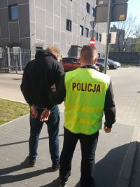 Na zdjęciu widzimy młodego mężczyznę obróconego tyłem do aparatu , ma założone kajdanki. Obok policjant w żółtej kamizelce policyjnej trzyma go za rękę. Chłopak ma na sobie czarną kurtkę.