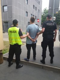 Na zdjęciu zrobionym na zewnątrz Komendy Miejskiej Policji widzimy młodego chłopaka w okularach , w szarej luźnej koszulce, odwróconego tyłem do aparatu. Ma założone z tyłu kajdanki. Po jego obu stronach widzimy dwóch policjantów , w czarnym umundurowaniu , jeden z nich ma żółtą kamizelkę z napisem Policja.