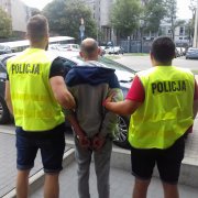 Na zdjęciu widoczni dwaj policjanci trzymający zatrzymanego. W tle parking policyjny. Osoby na zdjęciu ustawione są tyłem do obiektywu. Policjanci mają na sobie żółte kamizelki odblaskowe z napisem Policja.