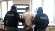 Na zdjęciu dwóch policjantów ubranych w czarne kamizelki taktyczne z napisem Policji. Policjanci trzymają zatrzymanego, który ma założone kajdanki trzymane na ręce złożone z tyłu. Osoby stoją w pomieszczeniu tyłem do obiektywu aparatu.